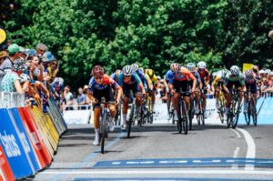 Group of competitive cyclists sprinting towards the finish line during the Santos Tour Down Under, with cheering spectators lined up along the barriers. The athletes are intensely focused, wearing brightly coloured team jerseys and helmets, with a backdrop of trees and a clear blue sky.